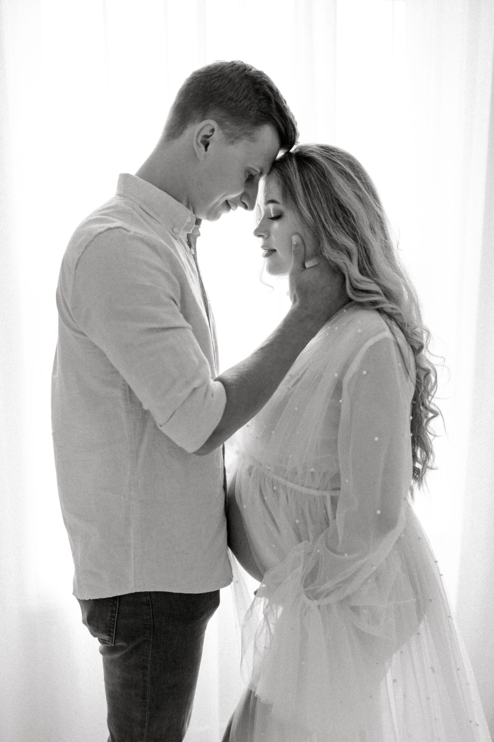 Black and white maternity portrait of a couple forehead to forehead by a window