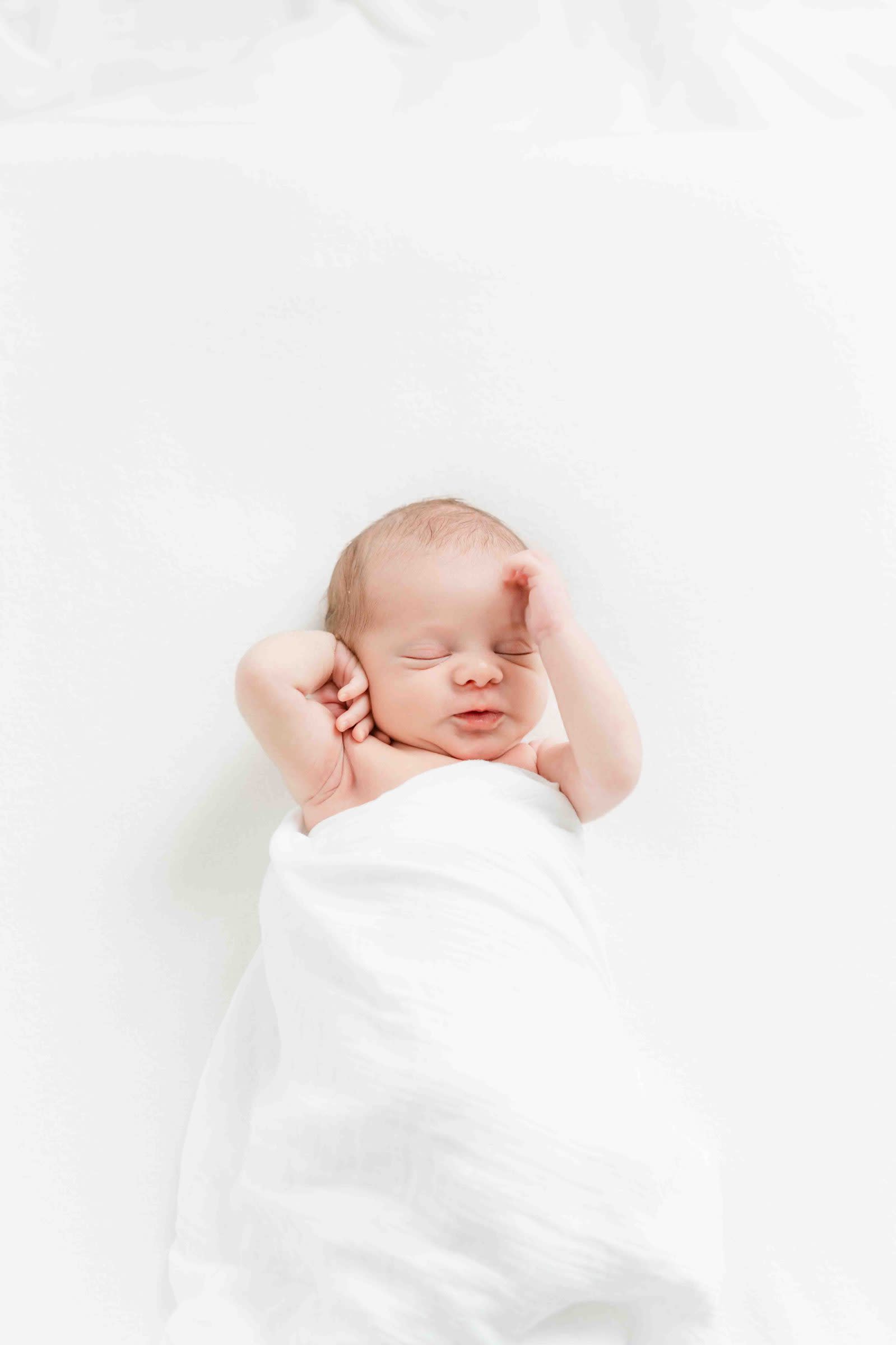 Newborn baby asleep in a white swaddle with hands raised near their face