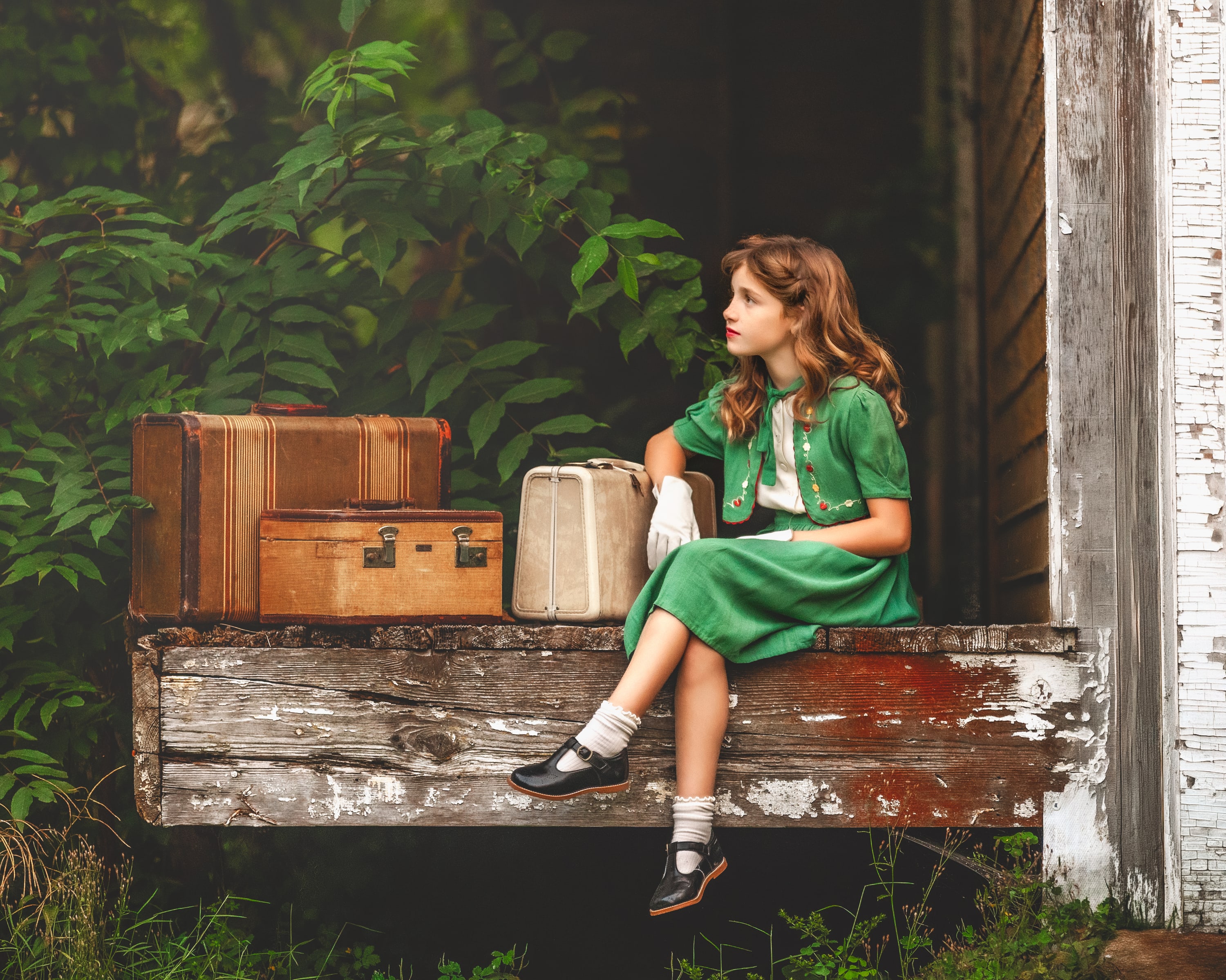 Young girl in a vintage green dress witting on the weathered edge of an old train station beside antique suitcases
