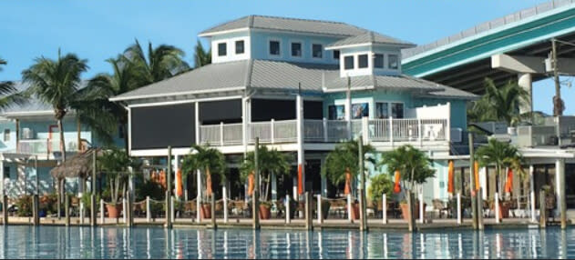 Waterfront restaurant with light blue building, balconies, palm trees, dock seating, and bridge in background.