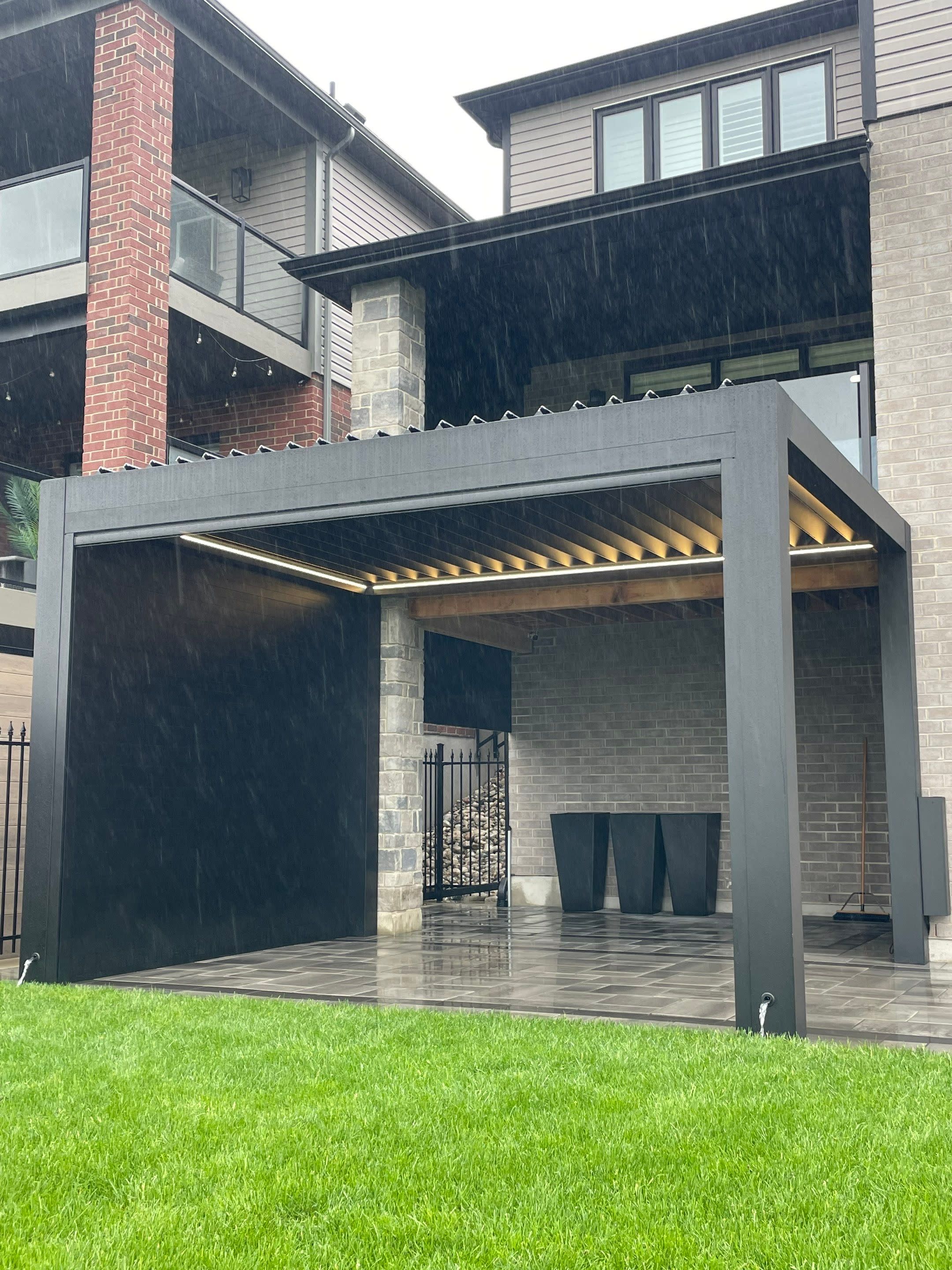 Umbra Gray aluminum louvered pergola with privacy wall and integrated drainage system during rainfall
