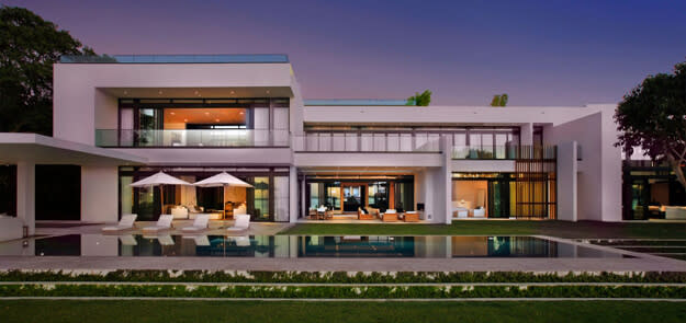 Modern white mansion at dusk with infinity pool, illuminated interior, and expansive glass walls.