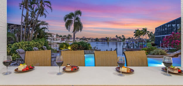 Outdoor dining counter with wine glasses and appetizers overlooking waterfront sunset with infinity pool.