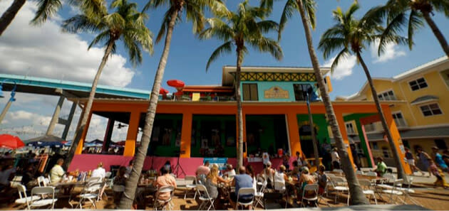 Outdoor patio restaurant with colorful building, tall palm trees, diners at tables under sunny sky.