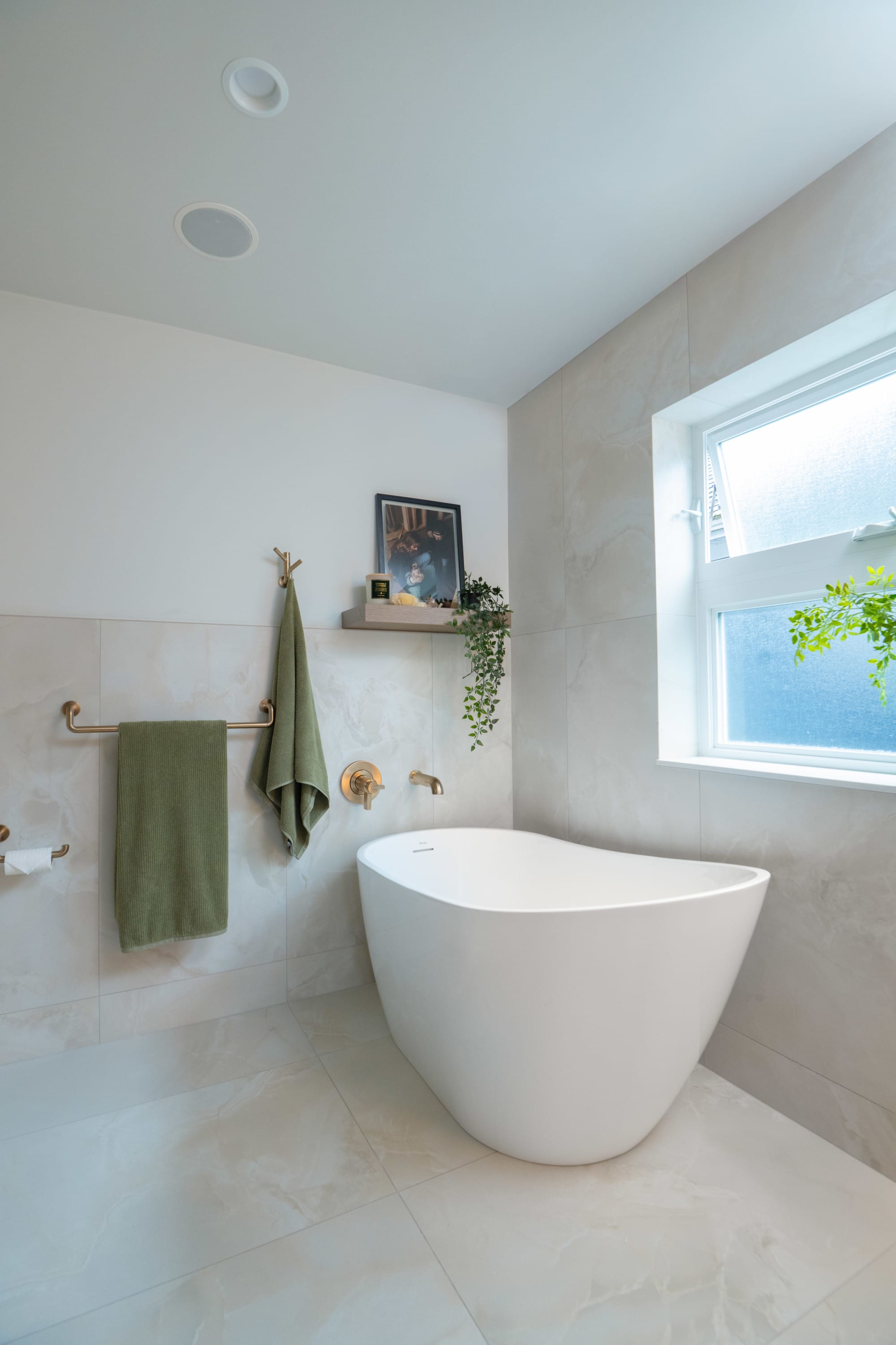 Bright modern bathroom featuring a freestanding white tub, soft neutral stone tile, brass fixtures, natural light, and minima
