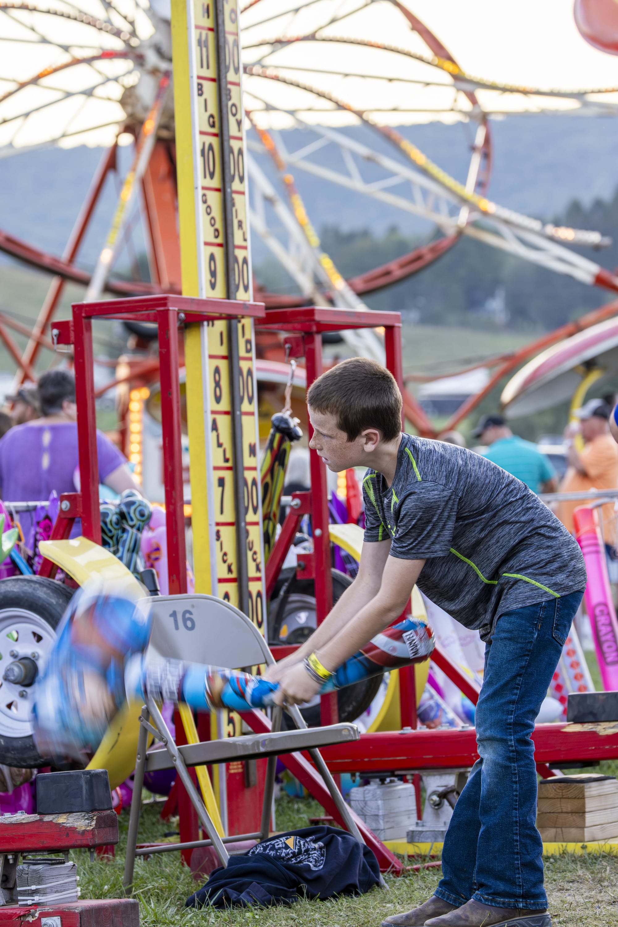 Person swinging a mallet at a carnival strength‑test game with rides and a Ferris wheel in the background