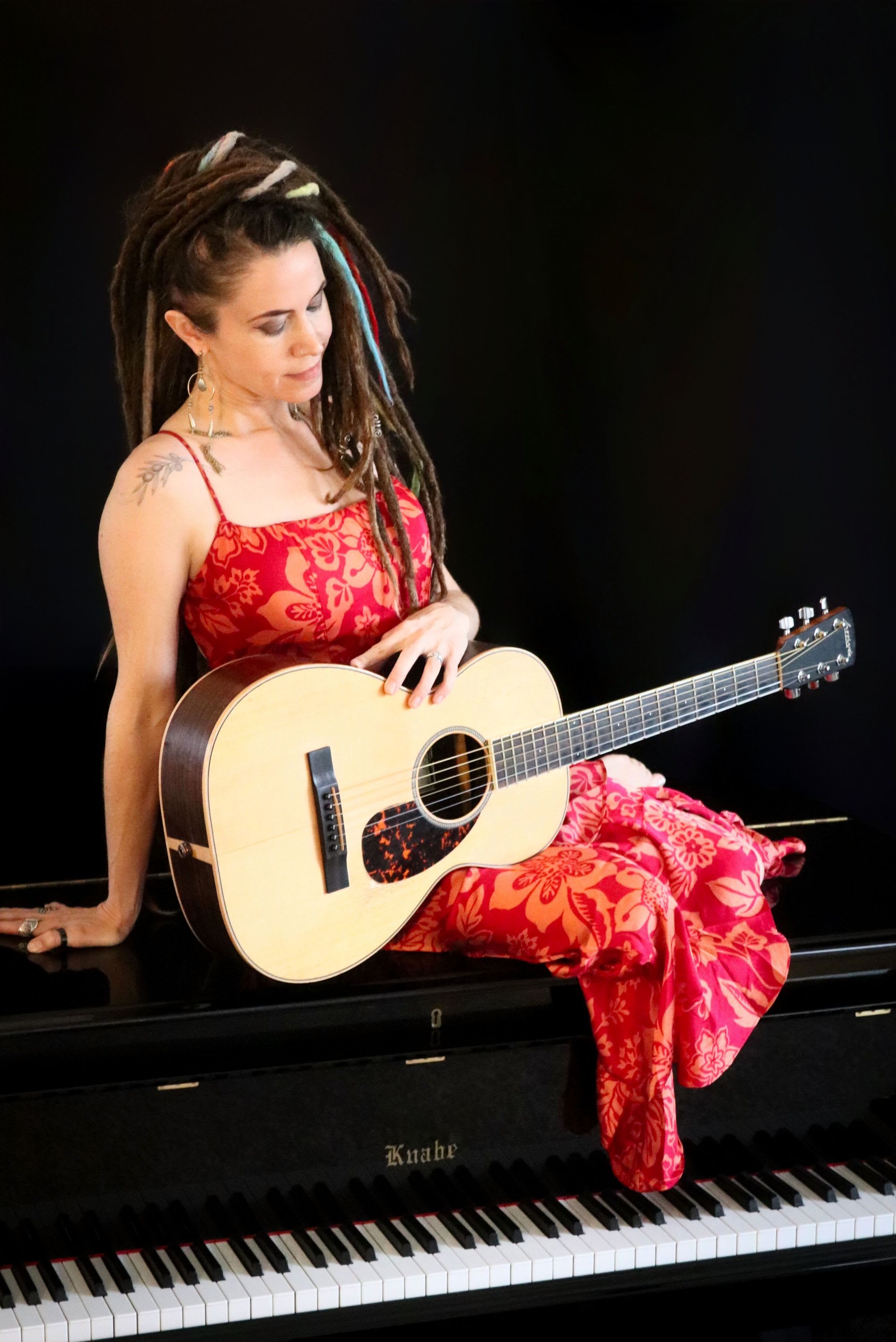 Woman in red dress with guitar sitting on top of a grand piano