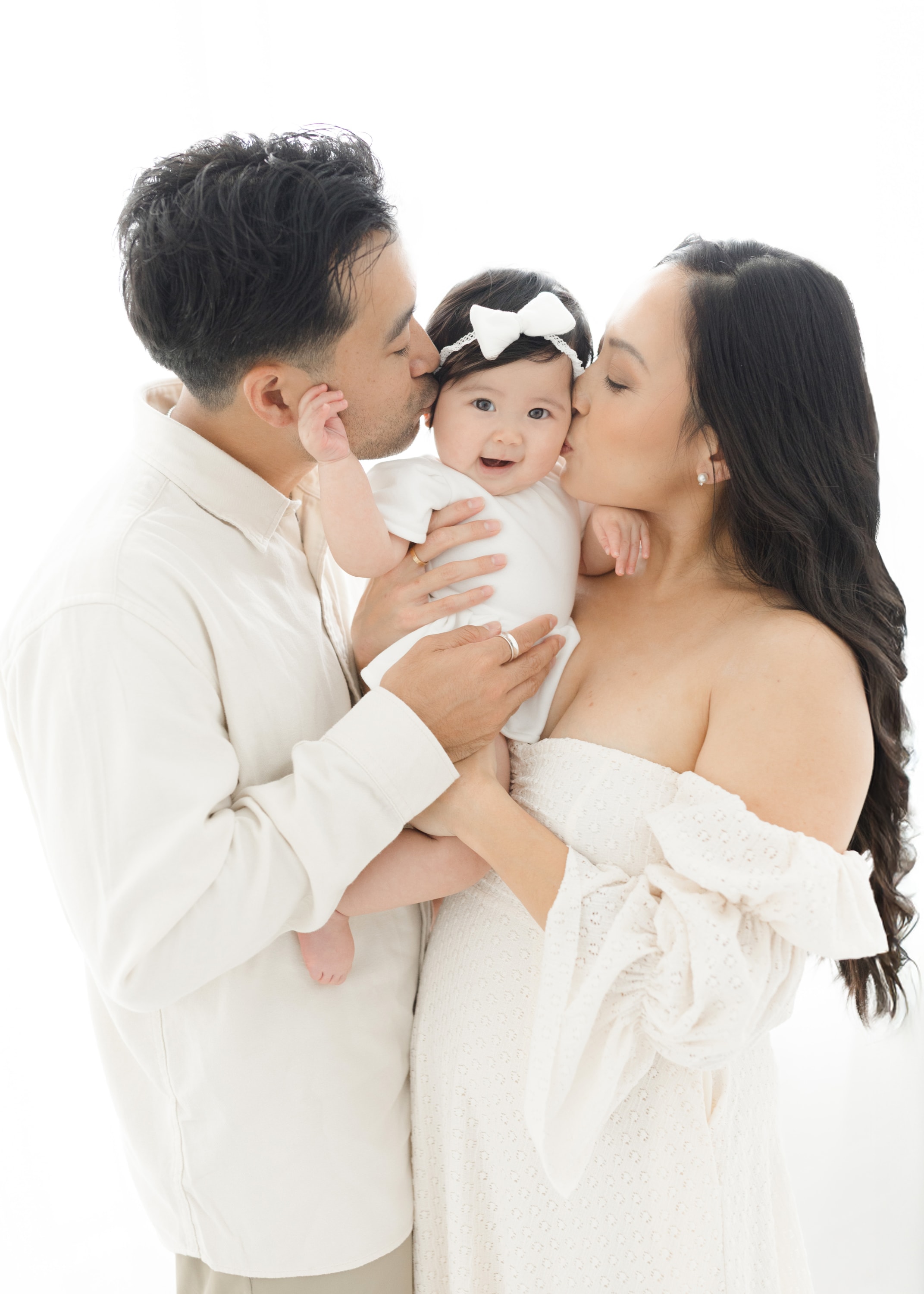 A mother and father gently kissing their baby girl on the cheeks standing in front of a window in a natural light studio