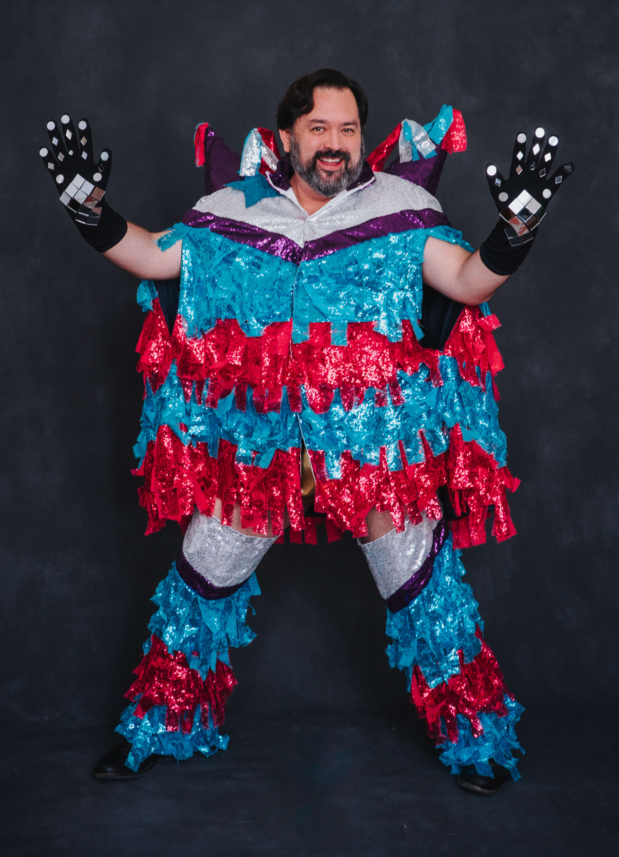 A dark-haired, light-skinned and bearded man is wearing a sparkly outfit with his hand pressed against the screen against a p