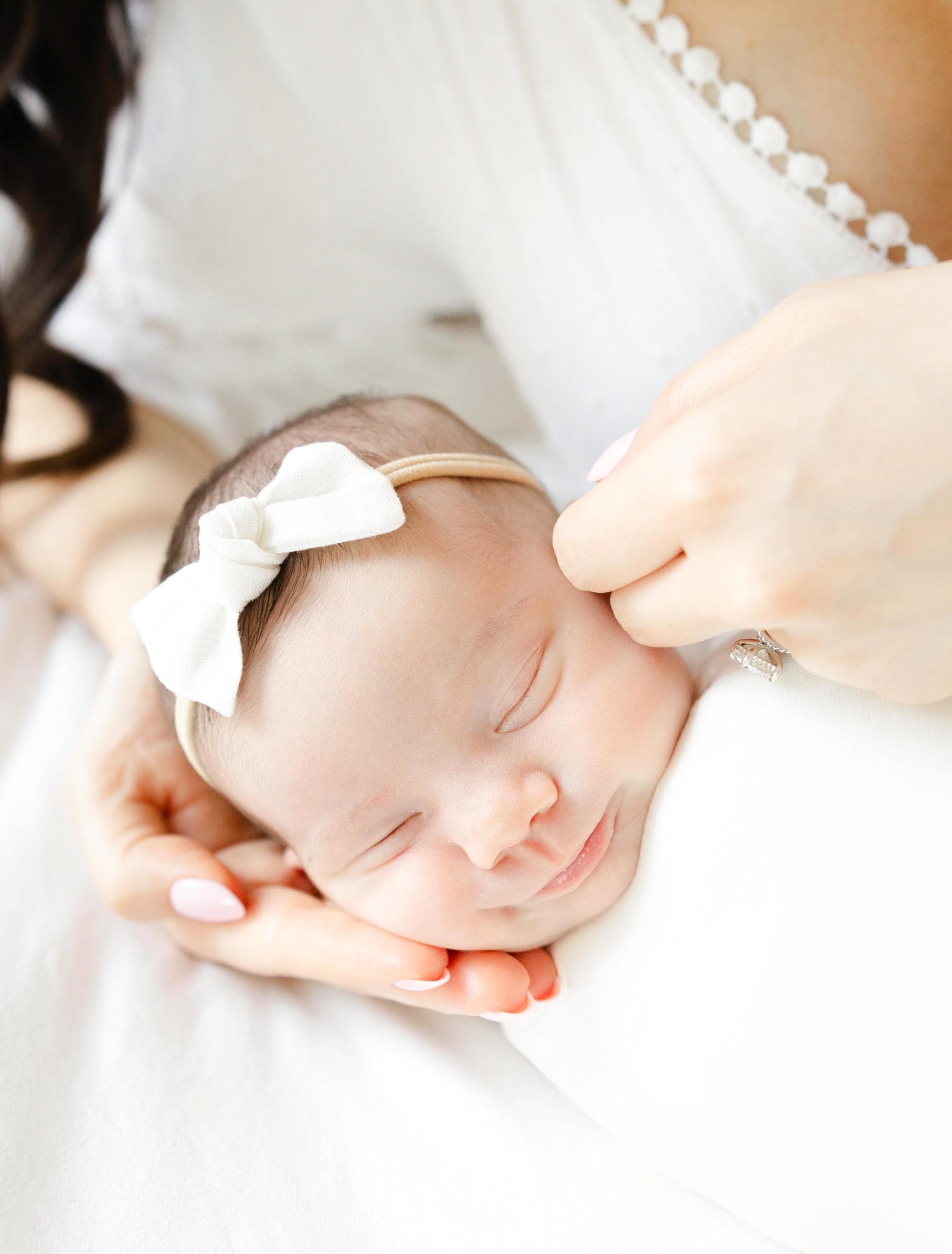 Sleeping newborn baby girl wearing a soft bow headband, gently cradled in a mother’s hands during a light and airy newborn ph