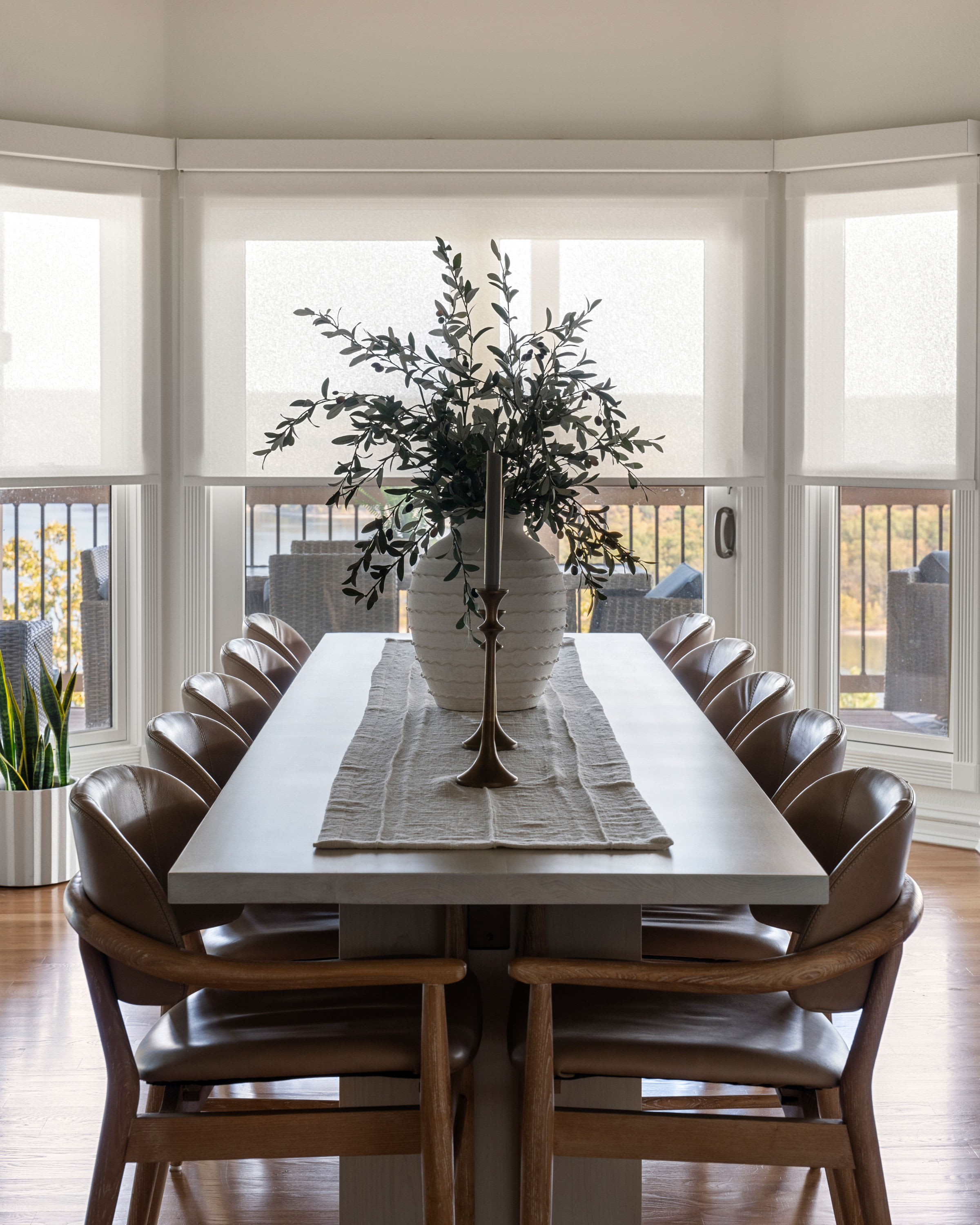 White light-filtering roller shades partially lowered across dining room windows behind a long table with brown leather chair