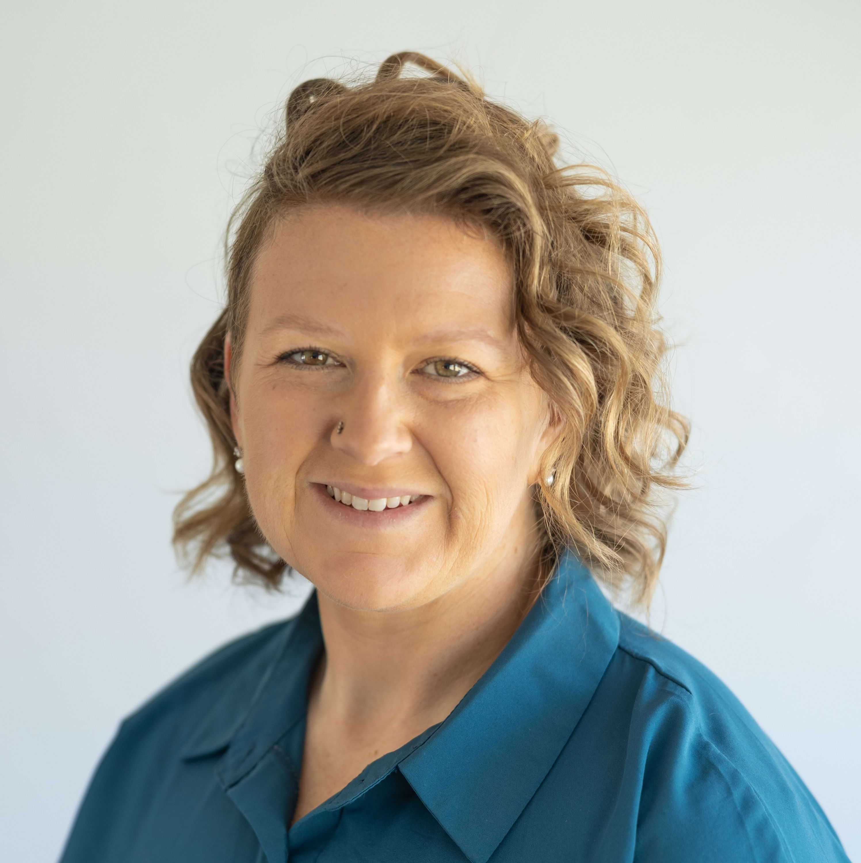 A close-up headshot of a smiling woman with wavy, honey-blonde hair wearing a teal collared button-down sh