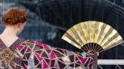 A performer in an ornate costume holds a gold folding fan against a modern architectural backdrop.