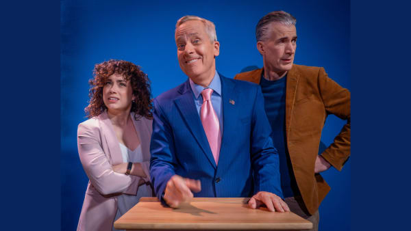 John Fisher, in a bright blue suit with American flag pin, mugs the camera. Elana Swartz and Gene Mocsy flank him, with quizz