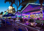 Restaurant patio at dusk with purple neon lights, palm trees, outdoor seating, and waterfront dining.