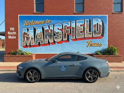 A 2024 Ford Mustang with the Aegis Cyber Defense logo parked in front of the iconic "Welcome to Mansfield" mural on a red bri