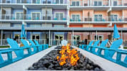 Outdoor resort seating area with turquoise chairs around a long fire pit in front of a colorful multi‑story building.