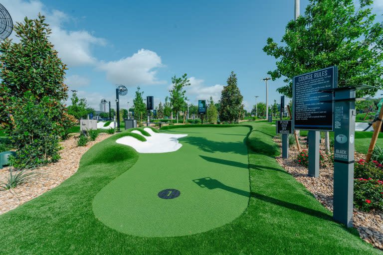 Mini golf course with bright green turf, curved putting areas, rocks, plants, and signs under a sunny sky.