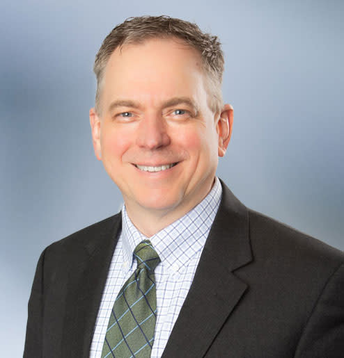 A middle-aged man, Thomas Huyck, smiles at camera, wearing a black sport coat with stripped patterned collared shirt and gre