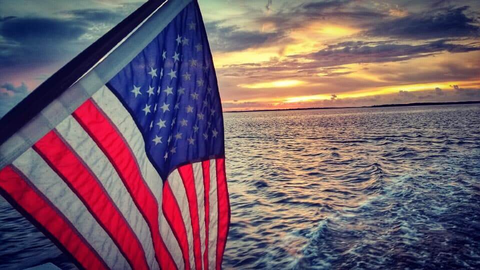 American flag waving in foreground with golden sunset over ocean waters behind it