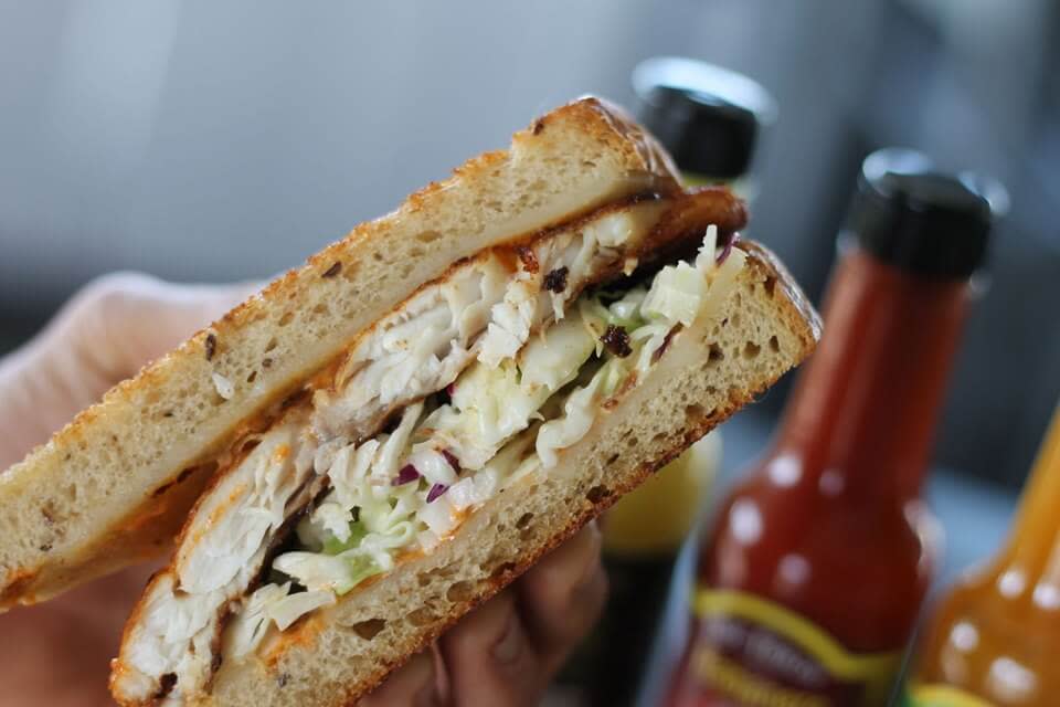 Grilled chicken sandwich on seeded bread with creamy coleslaw, hot sauce bottles blurred behind