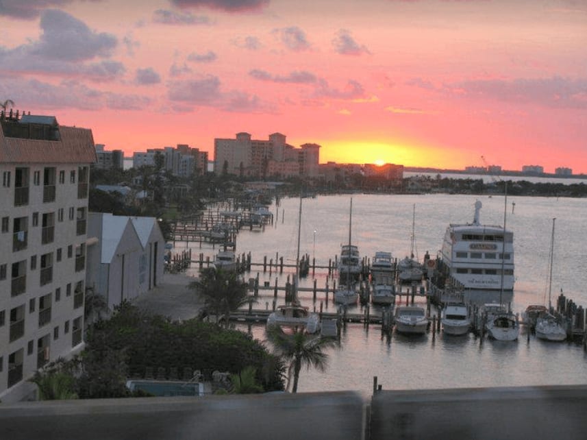 Marina at sunset with boats docked along waterways, buildings lining shore, and pink-orange sky.