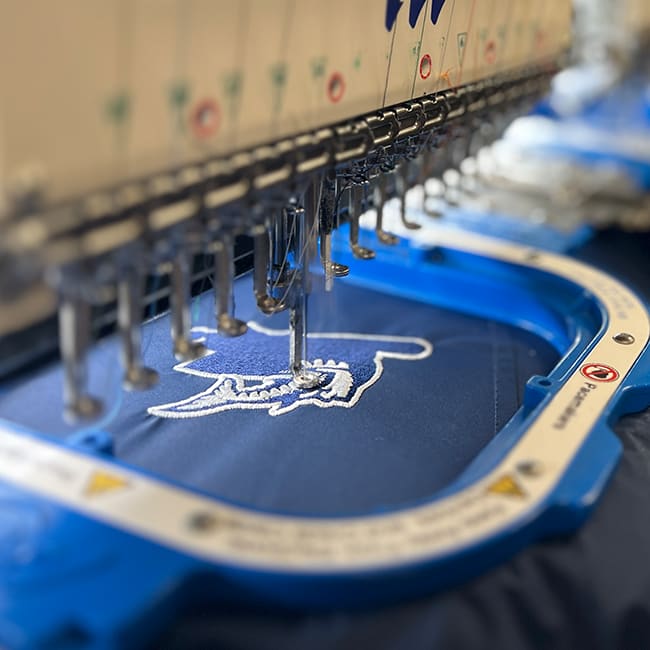 Close up photo of an embroidery production run showing white stitches on a blue garment
