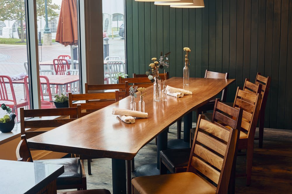 Long communal wood table set with linens and fresh flowers beside floor-to-ceiling windows overlooking the patio