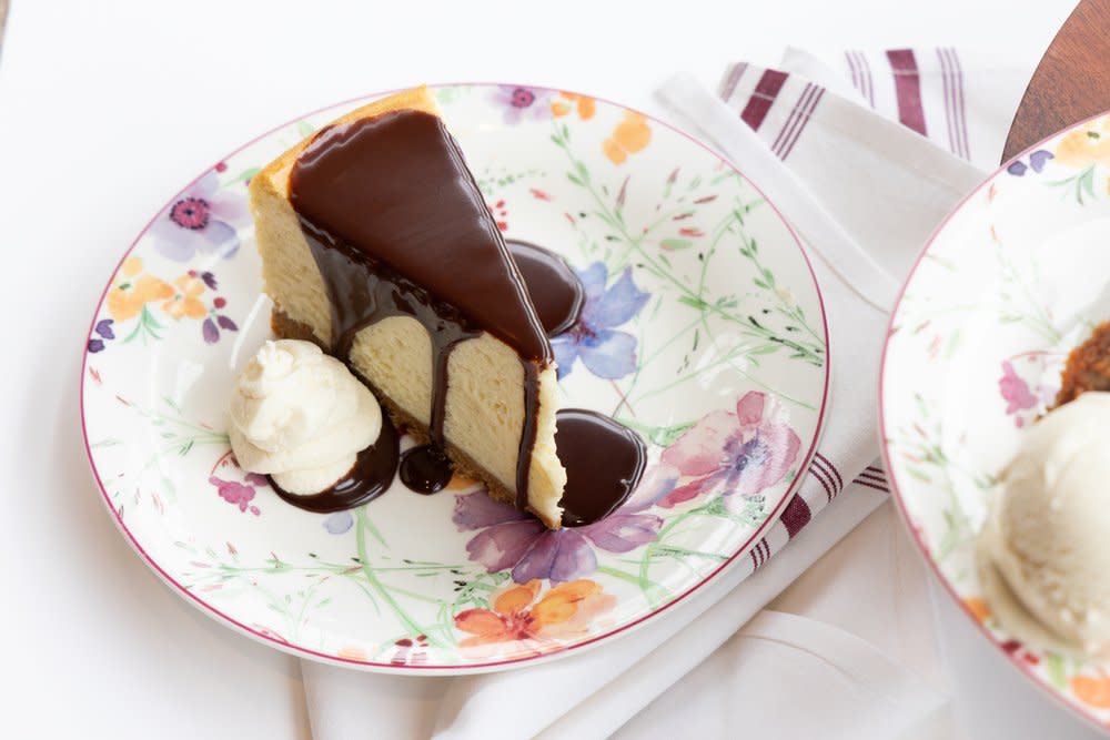 Slice of cheesecake covered in dark chocolate ganache served with a scoop of vanilla ice cream on a floral plate