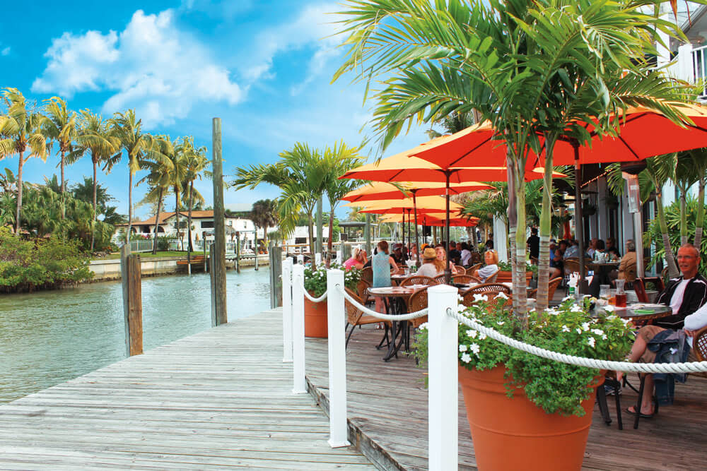 Wooden pier restaurant with orange umbrellas, palm trees, and diners along calm canal under blue sky.