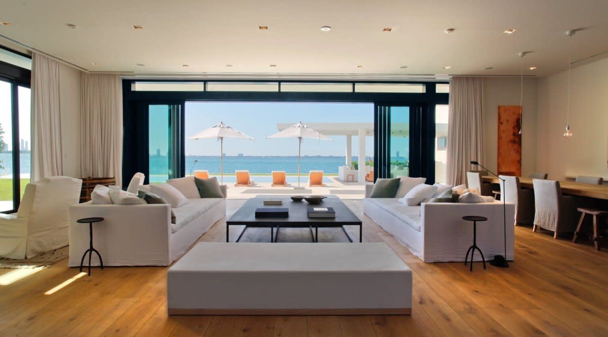 Beachfront living room with white sofas, floor-to-ceiling windows, and ocean view through glass doors.