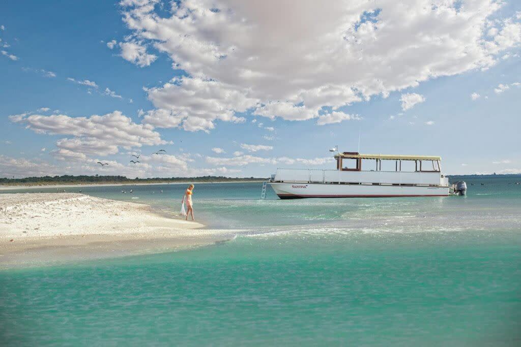 Person stands on sandbar near white tour boat in turquoise waters under cloudy sky