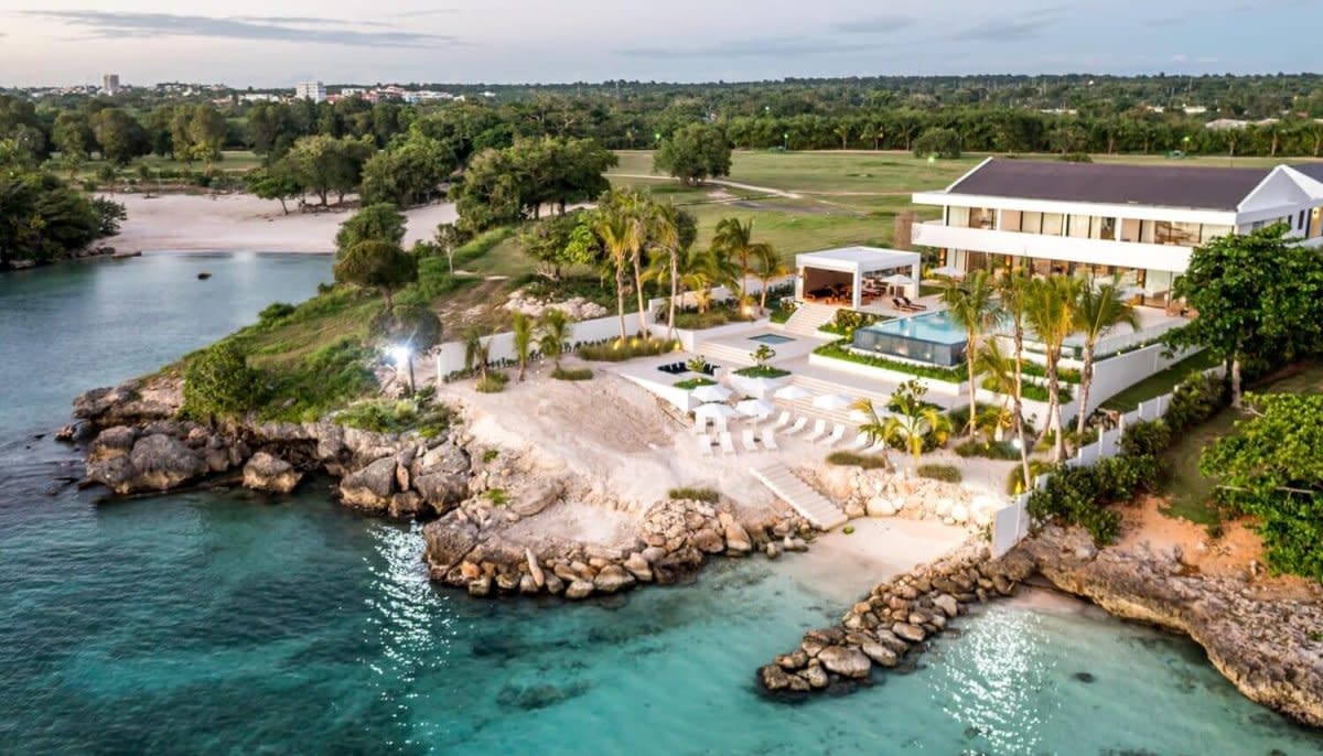 Aerial view of waterfront estate with white mansion, pool, private beach, and turquoise ocean waters.