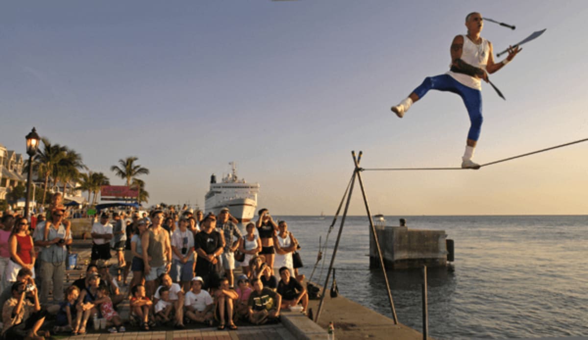 Street performer walks tightrope over water juggling knives while crowd watches with cruise ship background