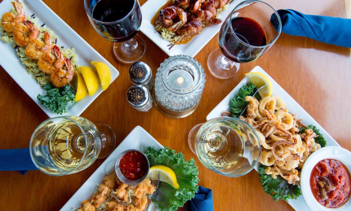 Seafood feast overhead view with shrimp, calamari, fish, wine glasses, and sauces on wooden table.