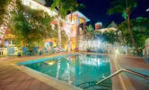 Resort swimming pool at night illuminated by building lights, surrounded by palm trees and patio seating