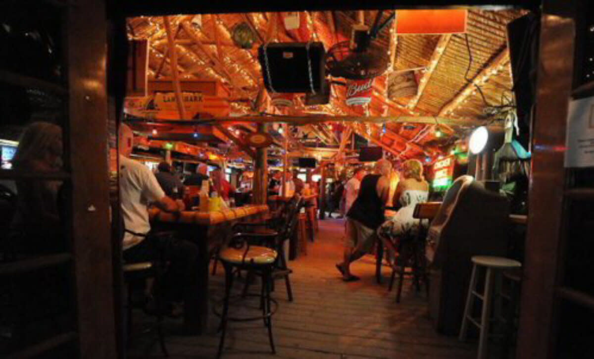 Warmly lit beach bar interior with wooden beams, patrons at bar counter and tables, rustic atmosphere