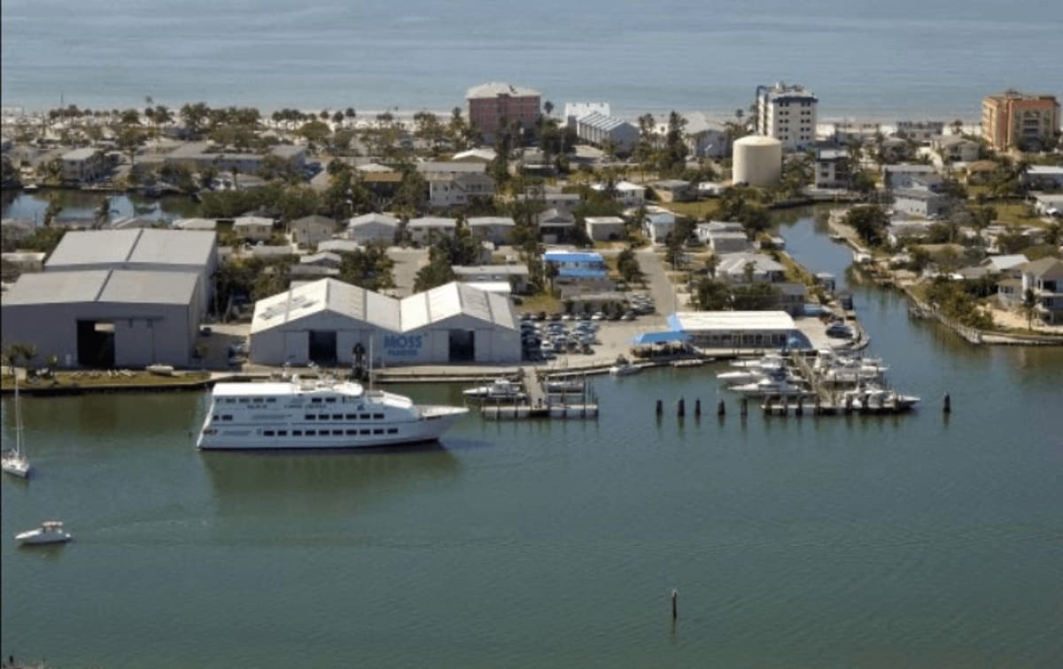 Aerial view of waterfront marina with white tour boat, storage buildings, and coastal community in background.