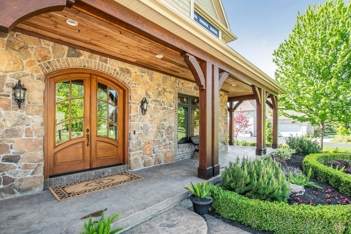 Custom home front entry with arched wood double doors, natural stone walls, and covered porch with timber columns by Spaces D
