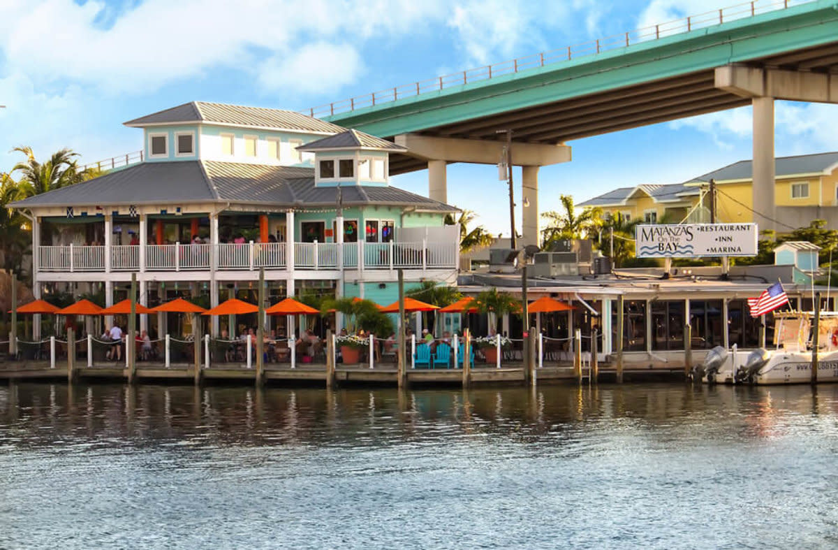 Waterfront restaurant with outdoor seating under orange umbrellas beneath bridge, American flag visible.