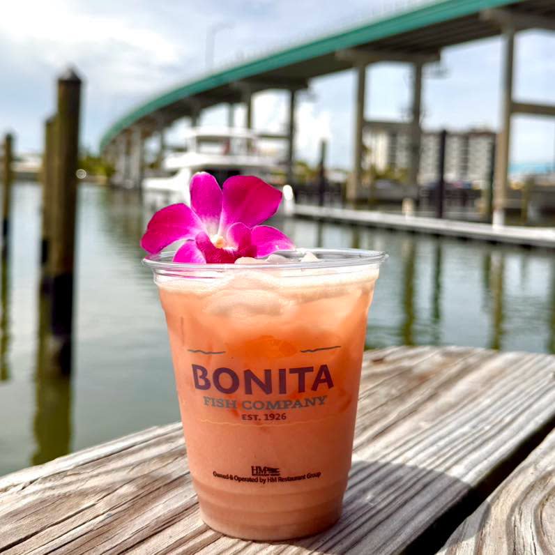 Tropical drink in a Bonita Fish Company cup garnished with an orchid, sitting on a wooden table overlooking a marina.