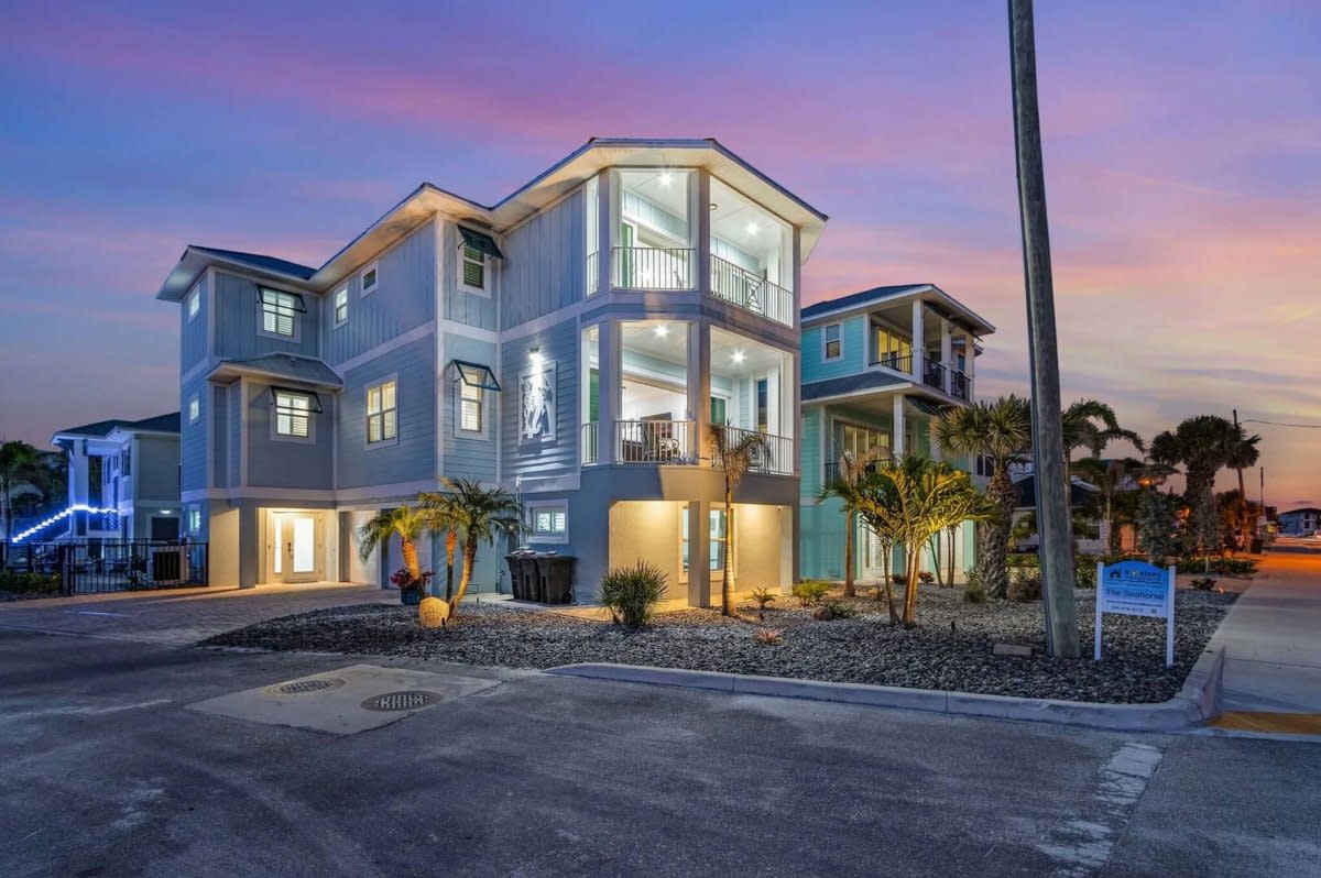 Three-story beach house exterior at sunset with illuminated windows, palm trees, and gravel landscaping.