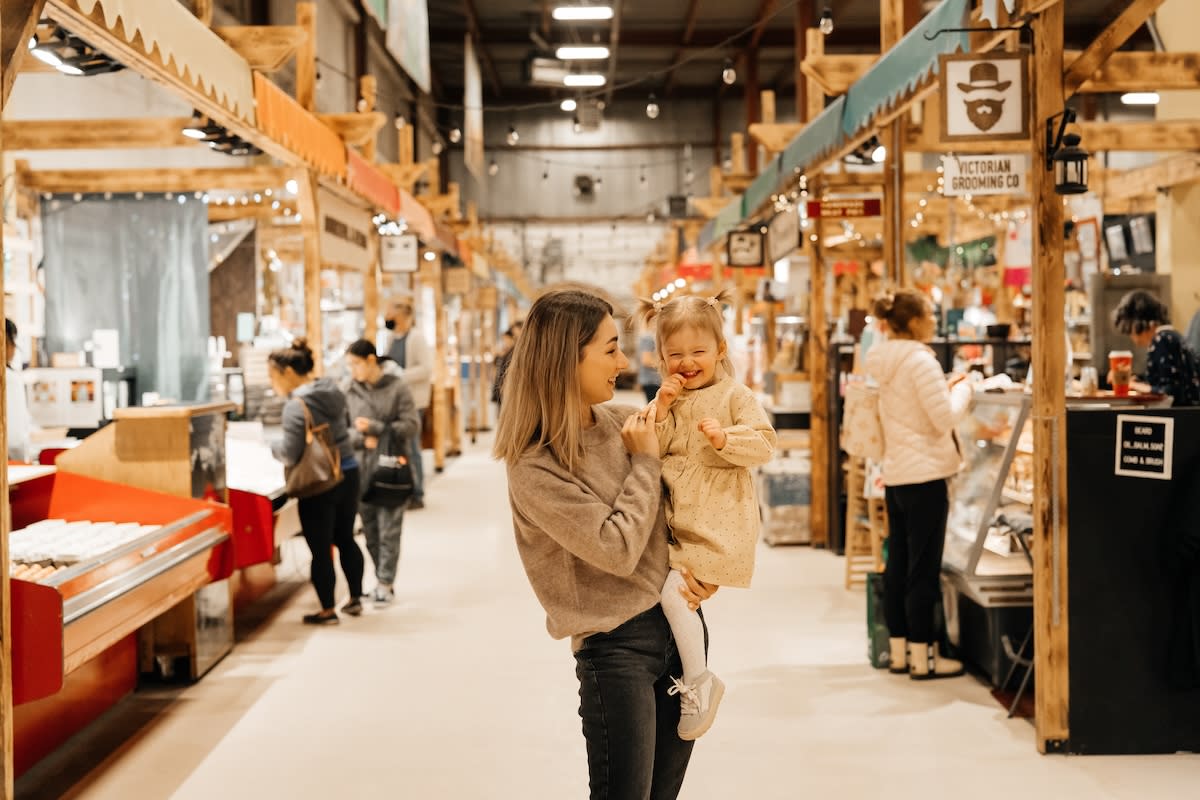 A mother holding her young child while walking through Bountiful Farmers’ Market, surrounded by local vendor stalls and shops