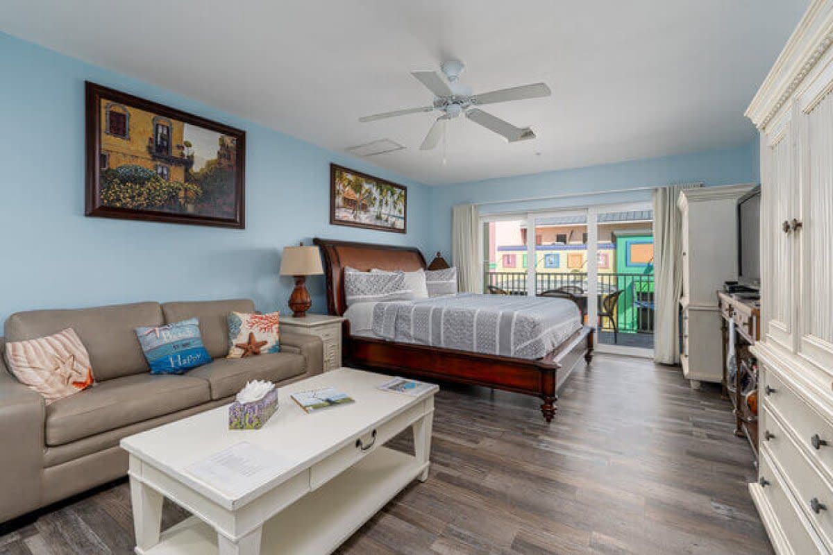 Open-plan studio with beige sofa, king bed, light blue walls, and balcony access