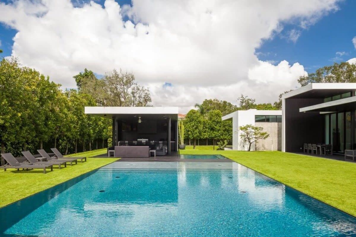 Long infinity pool between contemporary homes with green lawn, lounge chairs, and tropical trees.