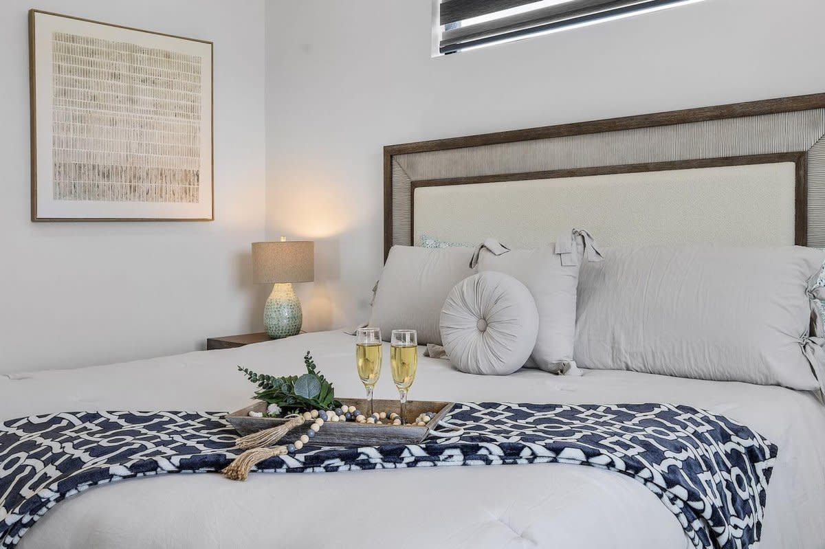 Bedroom with white linens, decorative throw, champagne glasses on tray, and framed wall art.
