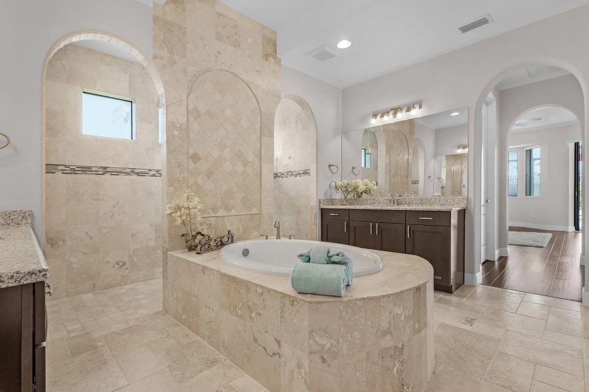 Luxury bathroom with oval soaking tub, travertine tile, dual vanities, and arched doorways.