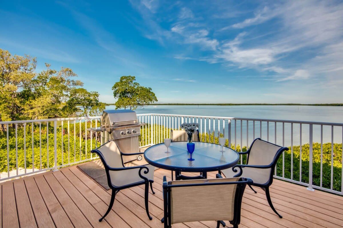 Wooden deck with glass table, four chairs, stainless grill, white railing, river view, sunny blue sky.