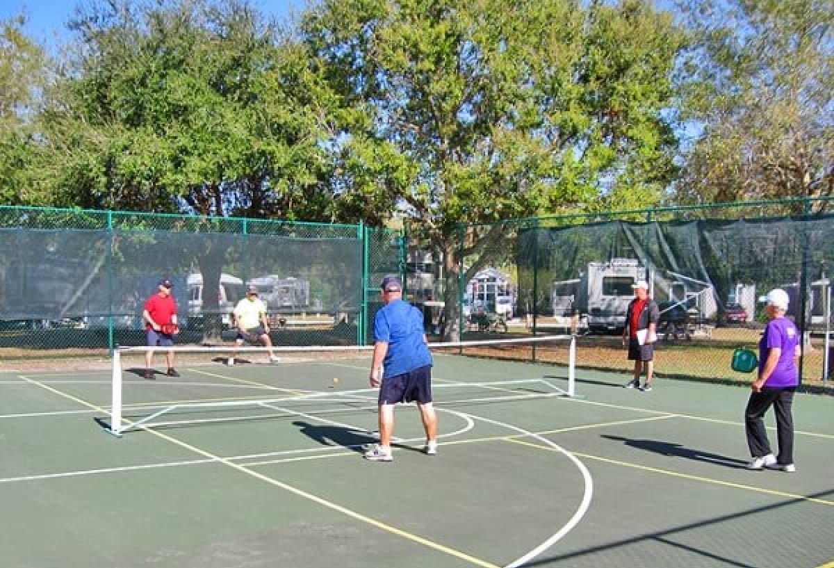 Adults playing pickleball on outdoor court surrounded by trees and campers