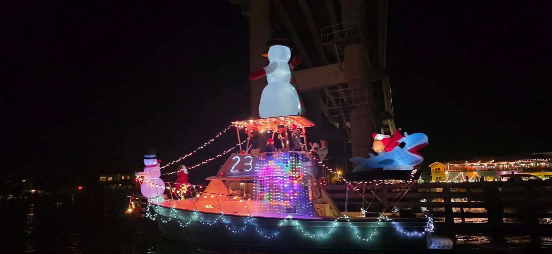 Night boat parade: vessel with glowing snowman, lights and "23" sign near a bridge